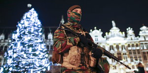 Vermummter Soldat mit umgehängtem Gewehr vor festlich beleuchtetem Weihnachtsbaum und Gebäude