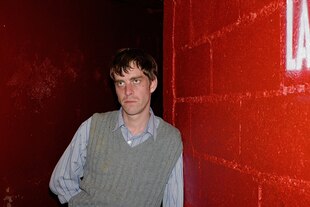 Saul Adamczewski von Insecure Men im grauen Pullunder lehnt an einer roten Wand