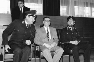 Otto Mühl mit Brille und Anzug sitzt zwischen zwei Polizeibeamten im Gericht. Er schaut ernst, während der linke Polizeibeamte mit Hut und Uniform einen gut gelaunten Eindruck macht