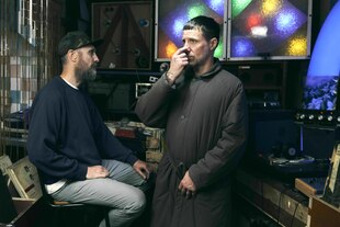 Die beiden Musiker stehen in einem Raum mit Schallplatten und Plattenspieler. Das Licht ist gedämpft - der Mann rechts im graugrünen, wattierten Mantel fasst sich an die Nase.Der Mann links sitzt auf einem Stuhl und schaut interessiert