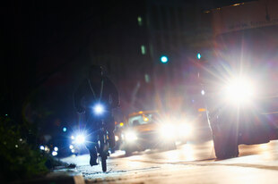 Fahrradfahrer im Dunkeln neben mehreren vorbeifahrenden Autos Fahrradfahrer im Dunkeln neben mehreren vorbeifahrenden Autos
