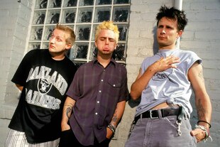 Die drei Mitglieder der Bank Green Day lehnen an einer Fassade mit Glasbausteinen - zwei versuchen cool rüberzukommen und der Mann in der Mitte mit blondiertem Haar macht Faxen