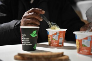 Auf einem Tisch stehen ein Becher Instantsuppe, aus der gelöffelt wird und ein Kaffeebecher. Auf einem Tisch stehen ein Becher Instantsuppe, aus der gelöffelt wird und ein Kaffeebecher.