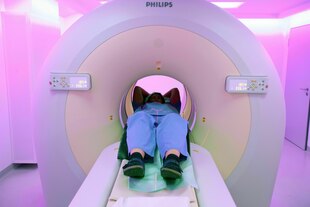 Eine Frau liegt in einer Röhre in der Radiologie, ihre Beine sind nicht in der Röhre. Sie trägt eine helle Hose und Birkenstock-Sandalen Eine Frau liegt in einer Röhre in der Radiologie, ihre Beine sind nicht in der Röhre. Sie trägt eine helle Hose und Birkenstock-Sandalen