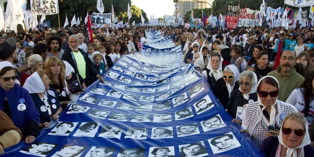 Viele Menschen auf einer Straße, die ein mehrere Meter langes Transparent tragen, auf dem Schwarz-Weiß-Fotos von Menschen sind - eine Gedenkveranstaltung für Opfer der argentinischen Militärdiktatur