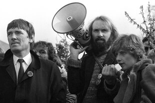 Otto Schily im Hemd mit Krawatte, Lukas Beckman mit Bart und langem Haar und Megafon in der Hand, in das Petra Kelly im Lammfellmantel, spricht