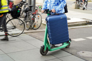 Eine Person fährt mit einem Koffer auf einem E-Scooter Eine Person fährt mit einem Koffer auf einem E-Scooter