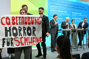 Ein Transparent mit der Aufschrift "CO2-Betäubung: Horror für Schweine" wird von einer Aktivistin auf einer Bühne in die Höhe gehalten, im Hintergrund stehen Vertreter aus Politik und Landwirtschaft. Ein Transparent mit der Aufschrift "CO2-Betäubung: Horror für Schweine" wird von einer Aktivistin auf einer Bühne in die Höhe gehalten, im Hintergrund stehen Vertreter aus Politik und Landwirtschaft.