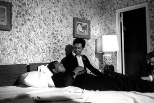 Das Schwarz-Weiß-Foto zeigt Muhammad Ali, der auf dem Hotelbett liegend mit Franz Beckenbauer spricht, der an der Bettkante sitzt. Auf der Blumentapete hängen Malereien in opulenten Bilderrahmen