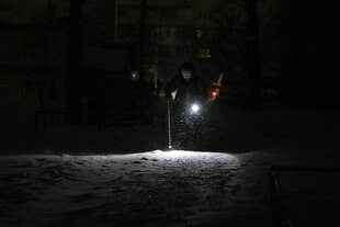 Eine ältere Frau, die mit einem Gehstock unterwegs ist, beleuchtet ihren Weg mit einer Taschenlampe auf einer Straße während eines Stromausfalls