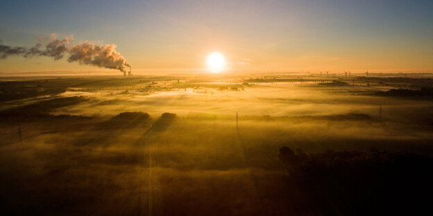 Bild von einem Sonnenaufgang, aus Kraftwerken steigt Rauch auf, Windräder sind zu sehen