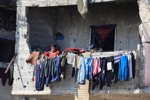 Zwei Frauen schauen über eine halbhohe Mauer, an der frisch gewaschene Wäsche hängt, die Wand ist voller Einschusslöcher , das Haus schwer beschädigt