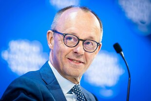 Friedrich Merz lächelt verschmitzt, er steht vor einer blauen Wand, neben ihm ein Mikrofon