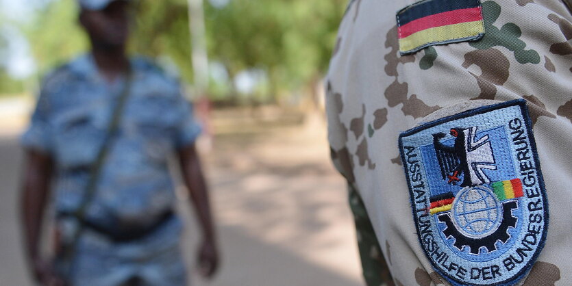 Ein Bundeswehrabzeichen an einer Tarnjacke ist scharf zu sehen, außer Fokus ist ein Mann, der im Hintergrund steht