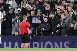 Ein Schiedsrichter sieht sich eine Szene am Bildschirm an, während das Publikum teils johlt, teils wütend schaut und ihn teils fotografiert. Ein Schiedsrichter sieht sich eine Szene am Bildschirm an, während das Publikum teils johlt, teils wütend schaut und ihn teils fotografiert.