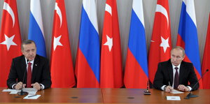 Erdoğan und Putin sitzen weit entfernt an einem Tisch, im Hintergrund Türkei- und Russlandfahnen