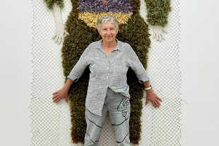 Eine Frau steht vor einer riesigen Frauenfigur aus Wolle
