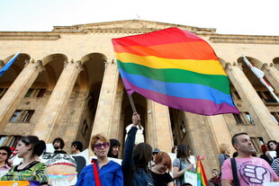Menschen bei einer Pride-Parade in Tiflis (Georgien) vor dem Parlamentsgebäude – bunte Regenbogenflaggen, Plakate und eine junge Person mit kurzem Haar hält eine große Regenbogenfahne hoch.