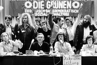 Schwarz-weiß Bild, 80er Jahre, im Hintergrund hängt ein Banner mit "Die Grünen" geschrieben, im Vordergrunde eine Gruppe Menschen, die meisten eher männlich gelesen. Vier davon Sitzen an einem Tisch mit dem Schild Diskussionsleitung