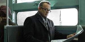 Tom Hank sitzt in der Bahn, hält eine Zeitung und schaut über den Rand seiner Brille