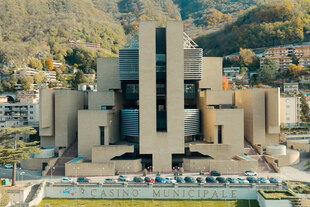 Das Bild zeigt den imposanten, modernistischen Neubau des Casinò di Campione von Mario Botta, der als monumentaler Block über dem Ort Campione d'Italia und dem Luganersee thront.