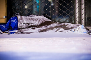 Eine obdachlose Person schläft bei winterlichen Bedingungen im Freien. Die Person liegt unter einer Decke vor einem Metallgitter; am Boden und auf der Decke liegt Schnee.