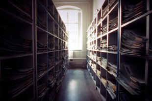 Blick in einen Flur zwischen zwei Regalen mit Alkten, Licht scheint frontal durch ein Fenster.