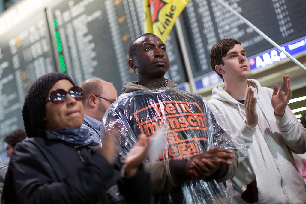 Drei Personen vor der Anzeigentafel des Frankfurter Flughafens, eine trägt ein T-shirt mit der Aufschrift "Kein Mensch ist illegal"