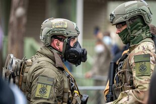 Zwei behelmte ICE-Beamte, der eine mit einer Gasmaske, stehen sich gegenüber