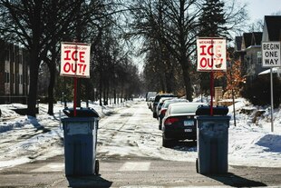 Mülltonnen stehen auf einer verschneiten Straße, an ihnen sind Protestschilder befestigt, auf denen steht:"Neighbours say ICE out"