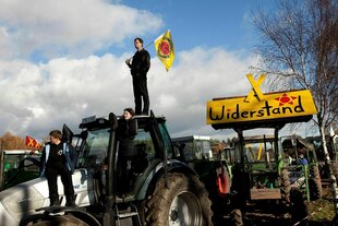 Ein Mann steht auf einem Traktor, er hat eine Fahne mit dem Logo der Anti-Atomkraftbewegung in der Hand, auf einem Transparent im Hintergrund ist das Wort "Widerstand" zu lesen