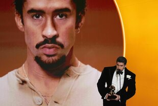 Bad Bunny steht im scharzen Smoking vor einem übergroßen Portrait seiner selbst, er hält einen Award in der Hand und spricht in ein Mikrofon Bad Bunny steht im scharzen Smoking vor einem übergroßen Portrait seiner selbst, er hält einen Award in der Hand und spricht in ein Mikrofon