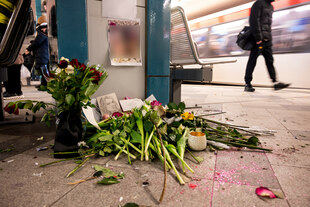 Eine Gedenkstätte mit Blumen, Kerzen und Zeichnungen auf dem Boden eines U-Bahnhofs, während im Hintergrund ein Zug vorbeifährt.