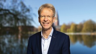 Porträt eines lächelnden Mannes mit Brille in einem dunklen Sakko vor einer unscharfen Kulisse aus einem Gewässer und herbstlichen Bäumen.