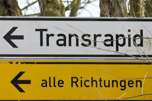 Zwei untereinander montierte Wegweiser, von denen der obere zum „Transrapid“ und der untere in „alle Richtungen“ weist.