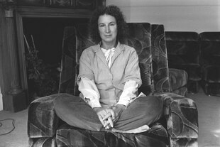 Schwarz Weiss Foto : Margret Atwood sitzt im Schneidersitz auf einem großen, dich gepolsterten Sessel und schaut ernst in die Kamera