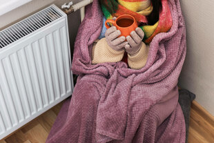 Eine Frau in Pullover, Handschuhen und Schal, eingewickelt in eine lila Decke, sitzt mit einer Tasse heißem Tee am Heizkörper Eine Frau in Pullover, Handschuhen und Schal, eingewickelt in eine lila Decke, sitzt mit einer Tasse heißem Tee am Heizkörper