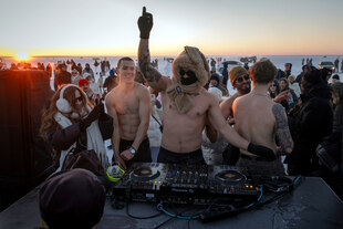 Männer stehen an einem Mischpult und tanzen mit nacktem Oberkörper in einer Schneelandschaft