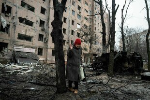 Eine Frau mit roter Mütze und Einkaufsbeuteln vor einem schwer zerstörtem Wohnhaus