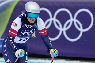 Lindsey Vonn mit Helm, Brille und Skianzug stützt sich auf ihre Stöcke, im Hintergrund die olympischen Ringe