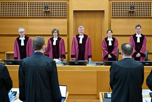 Fünf RichterInnen in roten Roben stehen hinter der Richterbank, vor ihnen im Vordergrund und der Kamera den Rücken zuwendend AnwältInnen in schwarzen Talaren