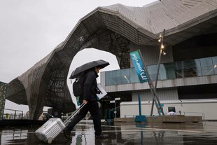 Ein Mann mit Schirm und Rollkoffer läuft auf ein großes, graues Betongebäude zu