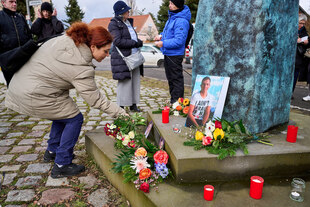 Eine Frau legt Blumen am Gedenkmal von Burak Bektaş nieder. Ein Foto von ihm lehnt an dem Denkmal, es liegen bereits Blumen davor und es sind Grabkerzen aufgestellt. Das Denkmal scheint von Menschen umringt, es sind durch Lücke in der dünnen Menschen Meng Eine Frau legt Blumen am Gedenkmal von Burak Bektaş nieder. Ein Foto von ihm lehnt an dem Denkmal, es liegen bereits Blumen davor und es sind Grabkerzen aufgestellt. Das Denkmal scheint von Menschen umringt, es sind durch Lücke in der dünnen Menschen Meng