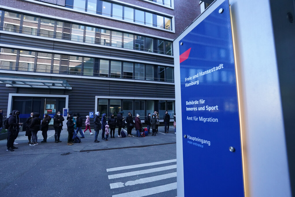 Im Dämmerlicht stehen Menschen Schlange vor einem Behördengebäude, auf einem Schild im Vordergrund ist