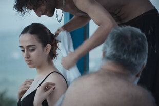 Im Vorderrgrund der behaarte Rücken eines älteren Mannes, dahinter beugt sich ein junger Mann über eine junge Frau