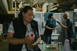 Sabine Thalau hält eine Flasche mit farbiger Flüssigkeit, im Hintergrund ein Mann und eine Frau in grüner Arbeitskleidung.