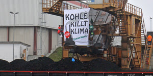 Kran mit Kohlehaufen davor, vor dem Steuerhäuschen hängt ein Plakat mit der Aufschrift "Kohle killt Klima", daneben zwei angeseilte Menschen