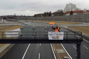 Ein Aktivist steht mit einem Schirm und einem Banner auf einer Schilderbrücke der A 100