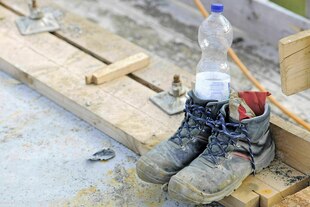 Dreckiger Arbeitsschuhe, in denen eine Flasche Wasser und ein Tuch stecken, stehen auf einem Brett - Baustellenambiente Dreckiger Arbeitsschuhe, in denen eine Flasche Wasser und ein Tuch stecken, stehen auf einem Brett - Baustellenambiente