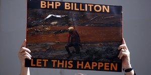mensch hält Plakat hoch, auf dem steht (deutsche Übersetzung): "BHP Billiton hat das zugelassen", auf dem Plakat ein roter Fluss zu sehen, alle Vegetation drumrum tot